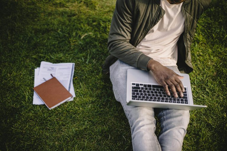 Crop black student with laptop and papers on green lawn